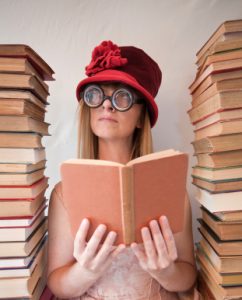 A woman wearing a red hat and glasses holds a book and sits between two stacks of books.