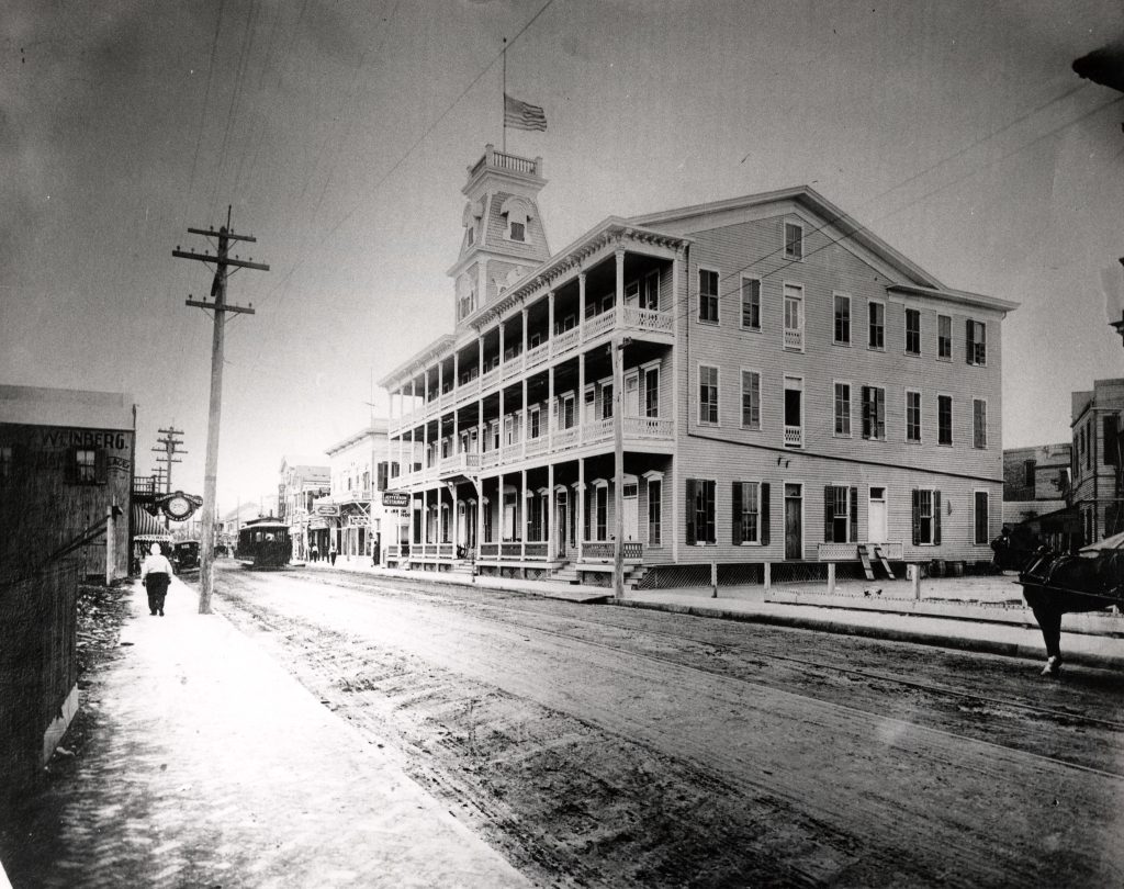 A black and white photograph with a three story wooden building with porches along the front facing the street, a person on the street, a street car and a pony in the front right hand corner.