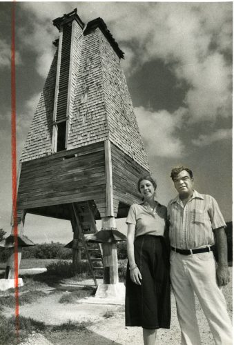 A woman and man stand in front of a shingled wooden tower.