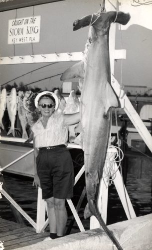 A woman stands next to a dead hammerhead shark hanging from ropes.