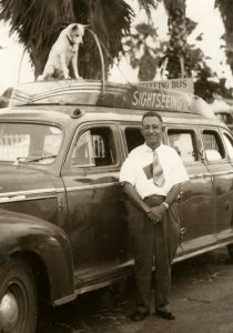 A man stands in front of a car with a sign on top that reads Sightseeing Bus and a dog on the roof looks down at him.