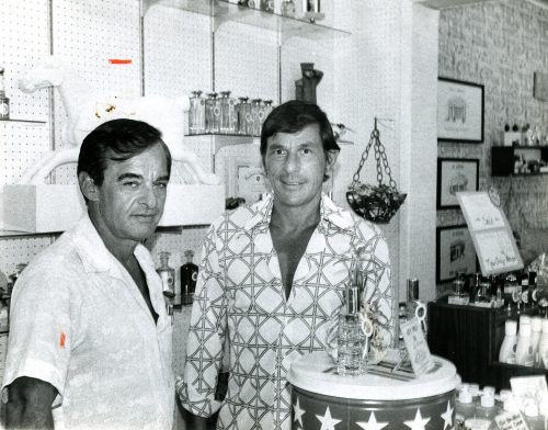 Two men stand behind a shop counter with clear bottles in front of them.