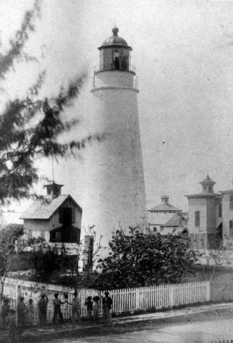 A row of people stand along a picket fence in front of a white lighthouse.