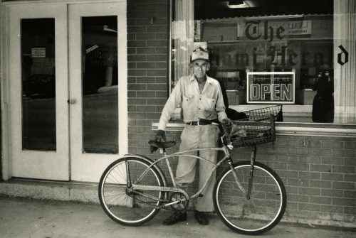 A man stands with a bicycle in front of an office with The miami Herald written on the window.