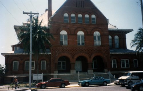 A three story red brick building with a chainlink fence and cars parked in front of it.