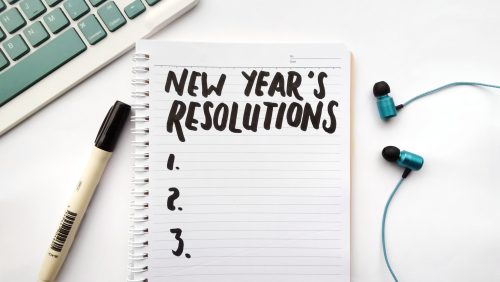 A pad of paper with New Year's Resolutions and the numbers one two and three, along side a pen, keysboard and earbuds.