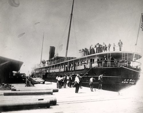 A steamship at a sock with people on both decks of the ship and standing on the dock.