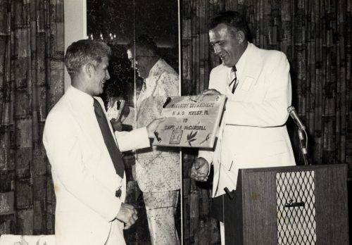 A man behind a lectern hands another man a plaque that says Security Department N.A. S. Key West FL to Capt. J.B. McCardell.
