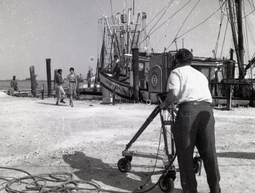 A man behind a camera films two people with a shrimp boat in the background.