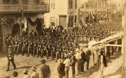 Rows of soldiers in uniform with rifles march down a street.
