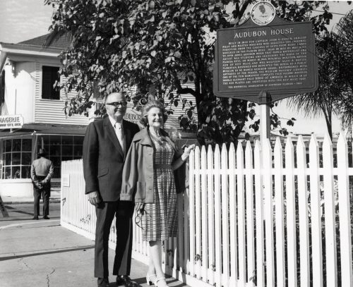 A man and woman stand in front of a picket fence and a sign that reads Audubon House.