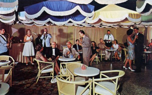 A band performs and a woman sings as people sit at small round patio tables under umbrellas.