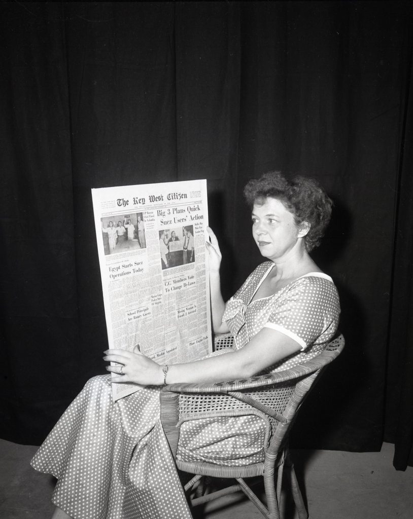 A woman sitting in a chair holds up the front page of a newspaper