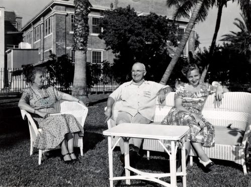 Three people sit on a sofa and chair on a lawn with building and palm trees in the background.