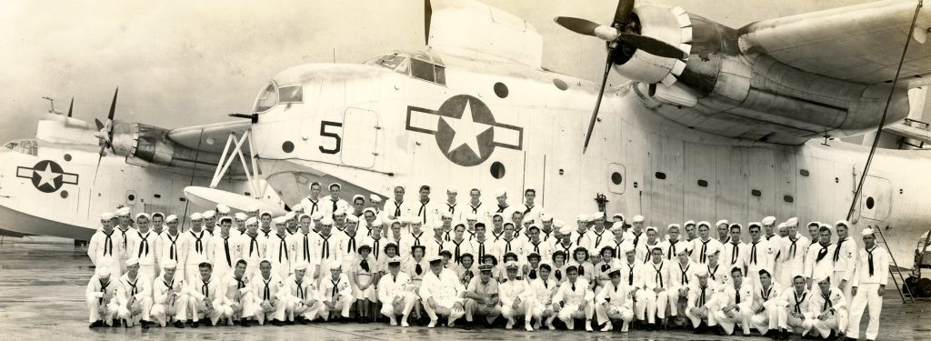 Three rows of Navy service members in white uniforms in front of a seaplane with a star on the side.