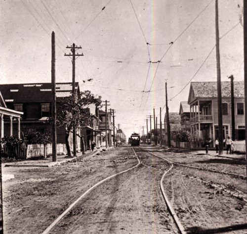 A dirt street with buildings on either side and trolley tracks in the middle with a trolley in the distance.