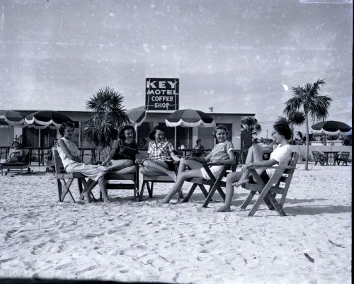 A group of women sitting on folding chairs on a beach in front of a building with a sign that reads Key Motel Coffee Shop.