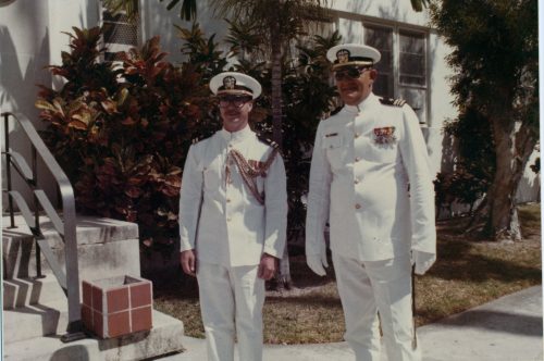 Two men in Navy dress uniforms stand in front of a white building