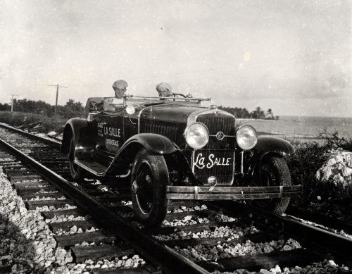 Two men in a 1920s car that is sitting on railroad tracks.