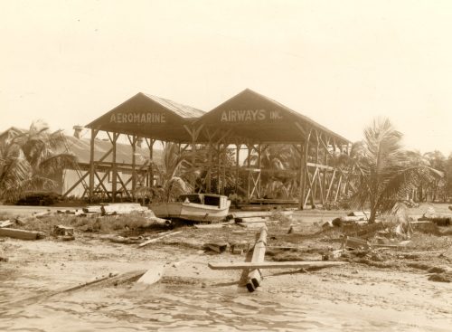 Two open sheds with the words Aeromarine Airways painted on them on a beach with wood and a boat hull in front of them.