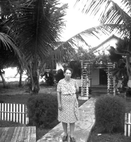 A woman stands on a walkway in front of a house with palm trees in the yard.