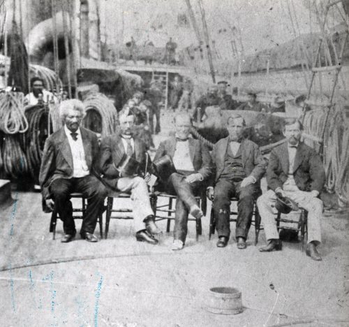 A row of five men sit on chairs on the deck of a ship.