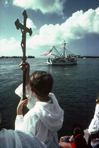 A child in a white robe holds a cross with a shrimp boat flying an American flag in the distance.