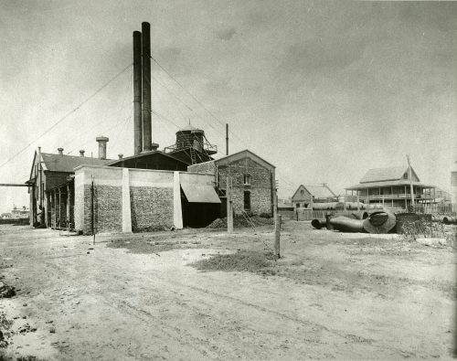 A brick building with two large smokestacks and houses in the background.