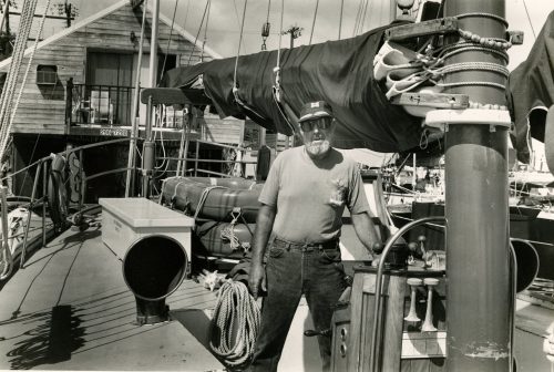 A man stands on the deck of a ship with a building behind him.