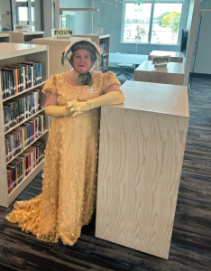 A woman in a yellow Regency-style dress and bonnet stands next to bookshelves.