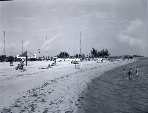A beach with antennas in the background and people on the beach and in the water.