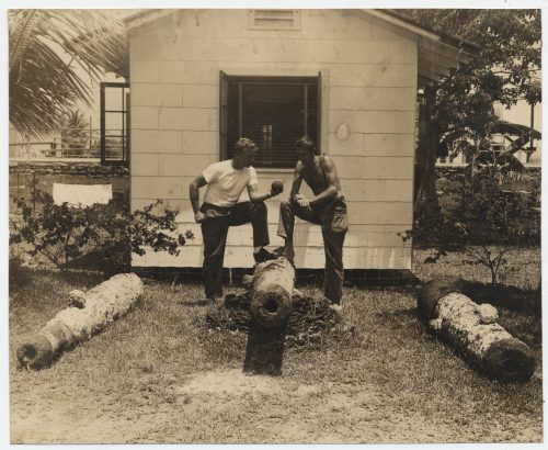 Two men standing in front of a building with three cannons on the ground, one of them holds a cannonball.