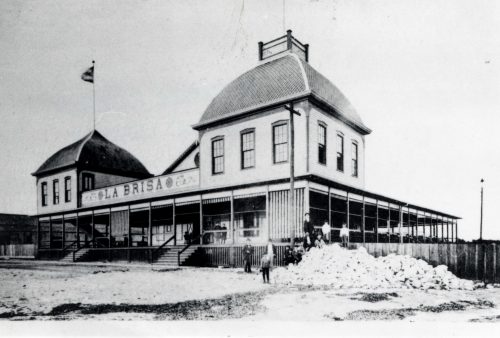 A building on the beach with two large cupolas, a wraparound porch and a sign that says La Brisa.