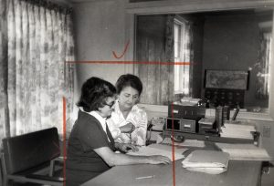 Two women sitting at a table examine a document. Photo has red wax lines and checkmarck.