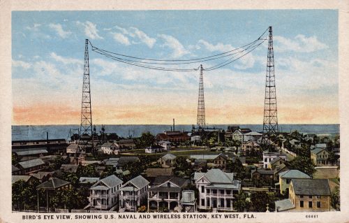 A colored postard showing buildings and towers connected by wires with water visible inthe background. Text reads Bird's Eye View, showing U.S. Naval and Wireless Station, Key West, Fla.