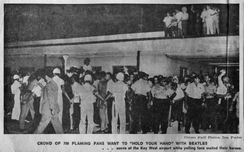 People standing in front of and on the roof of a building behind a police cordon. Caption reads Crowd of 700 flaming fans want to "hold your hand" with Beatles ... scene at the Key West airport while yelling fans waited their heroes. Citizen staff photos, Don Pinder.