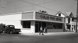 A man crosses a street and two women come out of a building with a sign that reads Carlos Food Center.