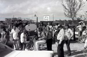 A group of people around a man sitting at a film camera with a sign in the background reading Cow Key Marina, Key West Florida.