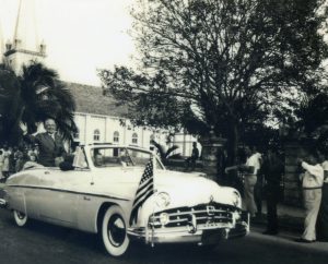 A man in a convertible with American flags on the front waves to people on the street.