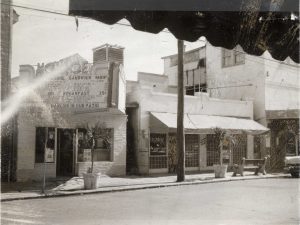 Several stores on a street front including one with a marquee that reads Monroe Sandwich Shop with a menu.