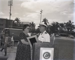 A man in a white Navy uniform hands a woman a plaque.